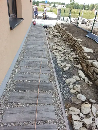 Úzký chodník z dlažby imitující dřevo podél domu, vedle kamenná zídka