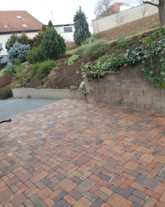 Dvůr z červenohnědé dlažby pod opěrnou zídkou se skalkou